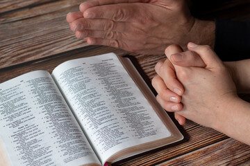 Naklejka premium Hands praying together in faith with an open Holy Bible Book on wooden background. Christian husband and wife pray and praise God Jesus Christ, a biblical concept.