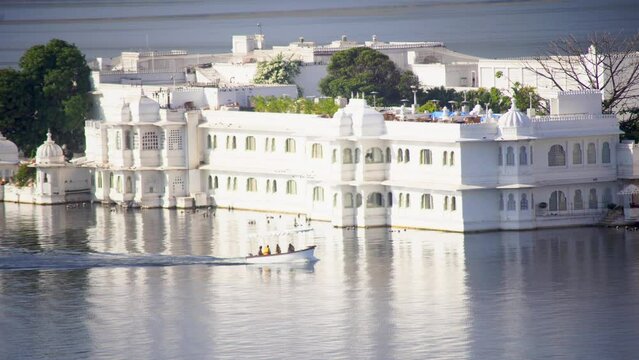 small tour boat with people crossing in front of city palace lake resort with ornate white building with rajput architecture of domes and arches on lake pichola in morning in udaipur rajasthan India