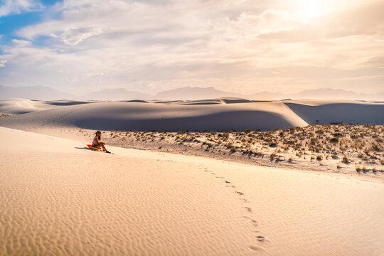 Young Woman Person Sitting On Famous White Sands Dunes National Monument In New Mexico On Disk Sled For Sliding Down Hill During Sunset With Vintage Brown Yellow Tone