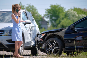Naklejka premium Stressed driver talking on sellphone on roadside near her smashed vehicle calling for emergency service help after car accident. Road safety and insurance concept