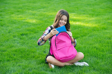 Fototapeta premium Happy teen girl taking book out of school bag sitting on grass. back to school. pupil at school time