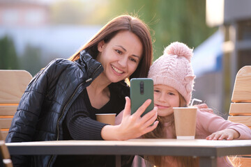 Young mother and her child daughter taking picture with phone selfie camera sitting at street cafe...