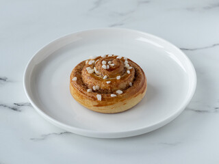 Traditional sweden cinnamon bun kanelbullar with cardamom sprinkled with pearl sugar. White plate on a marble table. Homemade pastry. Part of fika