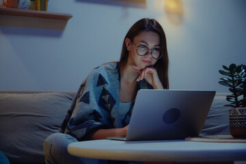 Young smiling woman using laptop at night, working overtime at home sitting on sofa, flexible...