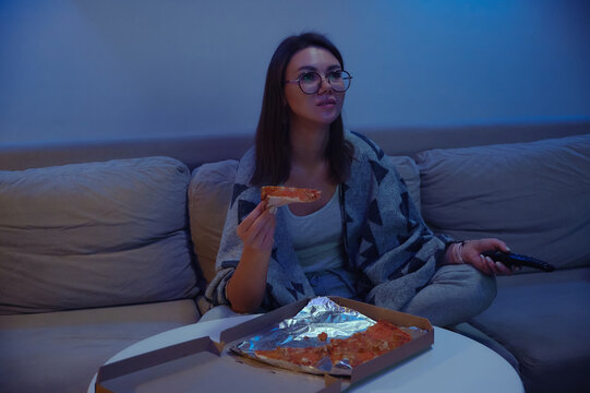 Happy woman eating pizza at night watching tv while sitting on sofa at home