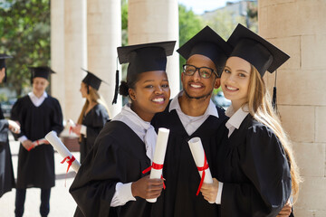 Portrait of three university graduate friends with diplomas in their hands outdoors after ceremony. Multiracial male and female students posing on campus background. Concept of international education