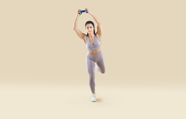 Fototapeta na wymiar Strong athletic woman trains with small dumbbell isolated on light beige background. Full length caucasian woman in sportswear set doing sports exercises. Concept of sports and active lifestyle.