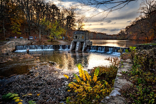 Morristown, NJ - USA - Nov 5, 2022 An Autumnal Horizontal Wide Angle View Of New Jersey's Historic Stone Speedwell Dam During Sunset. Fly Fisherman In The Foreground And People Hiking In The Distance.