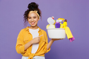 Young smiling happy fun housekeeper woman wear yellow shirt hold point index finger on basin with...