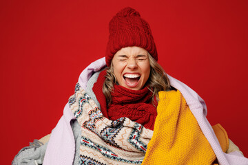 Young sad frozen woman wear scarf hat wrapped in many plaids close eyes scream shout isolated on plain red background studio portrait. Healthy lifestyle ill sick disease treatment cold season concept.
