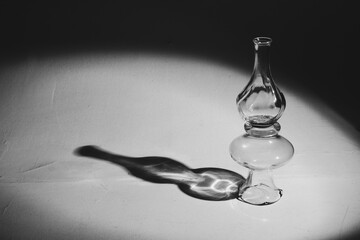 transparent crystal vase stands on a white surface. A light shines on top of the vase, creating a black silhouette shadow on the floor.