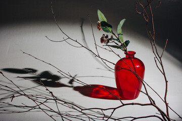 Still life with a red crystal vase with flowers inside and dead tree branches next to it on a white...