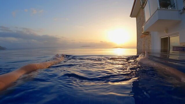 POV female hands rowing in water in infinity pool in paradise for relaxation and travel at sunset. girl swims in pool in blue transparent water, in background ocean with orange setting sun.