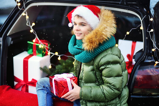 a child boy in a green knitted scarf and a Santa hat sits in the trunk of a car with a gift in his hands