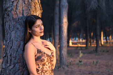 Fototapeta premium South American woman, young and beautiful, brunette, in leopard dress, leaning on the trunk of a tree, in the golden hour. Concept beauty, fashion, autumn, golden hour, melancholy.