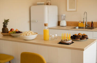 Kitchen with food and snacks prepared for a birthday celebration: orange juice, donuts, a cake with candles, fruit and popcorn.