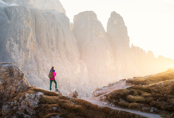 Young woman with backpack on the mountain trail and mountain canyon at sunset. Autumn in Dolomites, Italy. Colorful landscape with girl on path, rocks, golden sunlight in fall. Trekking and hiking