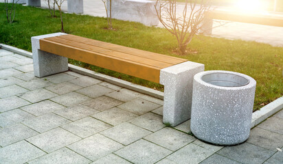 Empty concrete bench with wooden planks for sitting and garbage bin in city park with lawn and...