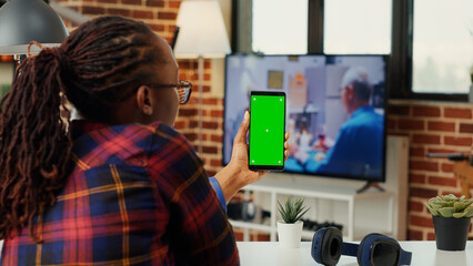 Young manager vertically holding mobile phone with greenscreen display, sitting at home office. Working with isolated chroma key template and blank copyspace, mock up background. Tripod shot.