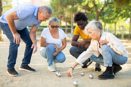 Mature white woman measuring distance between boules balls in petanque game outside in the park while other players gathered nearby 