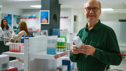 Senior customer smiling and looking at cardiology pills box in pharmacy, preparing to buy prescription medicine and pharmaceutical products. Checking supplements and medication on shelves.