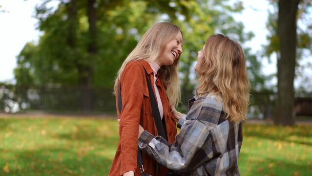 Side view two happy women hugging meeting in autumn park outdoors. Joyful young charming Caucasian friends talking laughing standing on alley outdoors. Friendship and lifestyle concept