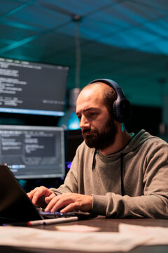 Young app developer writing code and listening to music on headphones, using artificial intelligence script to program big data on laptop. Programming server on terminal window.