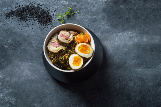 Sopa ramen, porchetta y huevo en taz&oacute;n blanco, sobre un plato negro y a un lado semillas de s&eacute;samo en un fondo oscuro	
