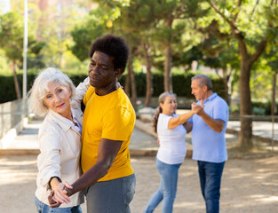 Two couples of various aged mixed-race opposite sex adult people dancing in pairs in public park on sunny day and having fun together