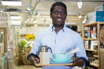Fototapeta na wymiar Satisfied african american male shopper holding dishware and kitchen accessories bought in household goods shop