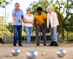 A group of multiracial middle aged mixed-sex adult people playing petanque game outdoors in public park on a sunny day