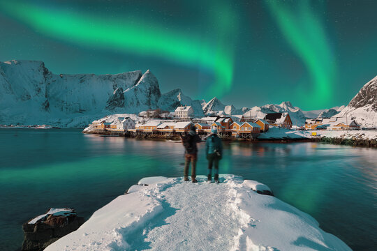 Сouple of tourists admire the view of the Sakrisoy village and snowy mountaines on background with Northern Lights