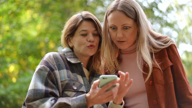 Beautiful Caucasian women scrolling social media in smartphone app gossiping in slow motion. Two relaxed confident young friends talking discussing rumors standing in autumn park outdoors