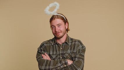 Portrait of smiling shy angelic young man with angel halo nimb over head flirting, looking at camera, positive emotions, celebrating holiday. Young blonde guy boy isolated on beige studio background - obrazy, fototapety, plakaty