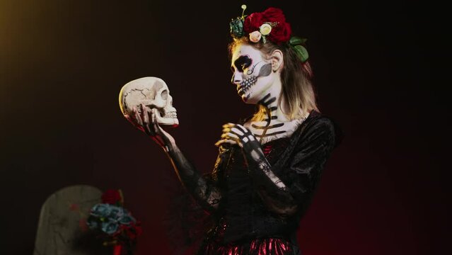 Holy goddess of dead talking to skull in studio, acting scary and horror to celebrate mexican halloween tradition. Santa muerte dressed in creepy festival costume with body art. Handheld shot.