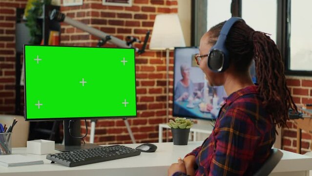 African american woman working with greenscreen and listening to song on headphones, using computer with chroma key display and isolated mockup. Looking at blank green screen.