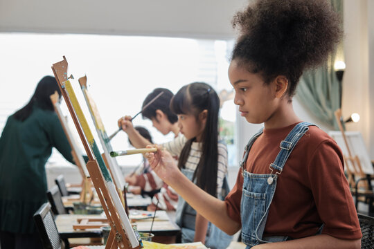 African American girl concentrates on acrylic color picture painting on canvas with students group in art classroom, creative learning with talents and skills in elementary school studio education.
