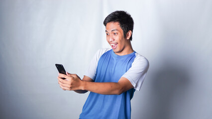 asian guy wearing tshirt looks suprised at the good news he received from his smartphone isolated white background