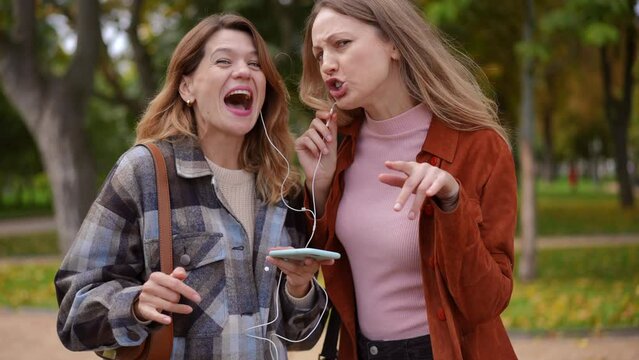 Portrait of two joyful women singing grimacing looking at camera enjoying hobby in autumn park. Front view of cheerful Caucasian friends posing having fun outdoors on weekend listening to music