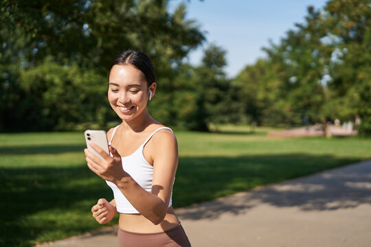 Happy Asian Fitness Girl Running And Checking Her Stats, Daily Goals On Smartphone App. Young Woman Jogging And Looking At Mobile Screen