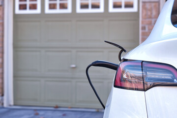 Back view of white EV car charging on driveway in front of garage at home with charger cable connected. Close up. Green energy and environmental concept.  