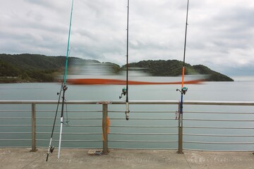 Fishing rods on the pier grid. Defocused ship passing through the harbor channel and the mountains behind. Santos city, Brazil.