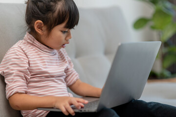Asian Little school kid girl use laptop computer sitting on sofa alone at home. Child learning reading online social media content, play education lessons game chatting with friends. 