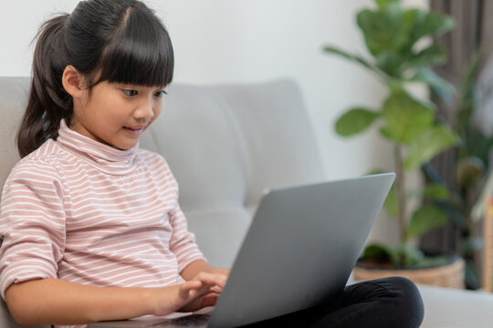 Asian Little school kid girl use laptop computer sitting on sofa alone at home. Child learning reading online social media content, play education lessons game chatting with friends. 