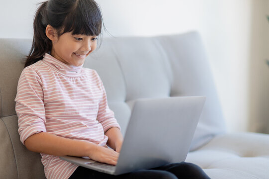 Asian Little school kid girl use laptop computer sitting on sofa alone at home. Child learning reading online social media content, play education lessons game chatting with friends. 