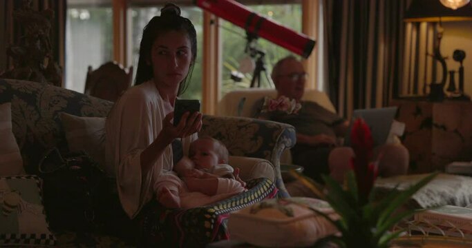 Mother with young daughter sits in living room with father in law behind her - family life - wide shot