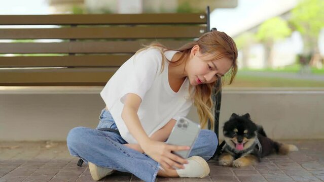 Happy young asian woman sitting on a bench and photo selfie in the park with her dog. Pet lover concept