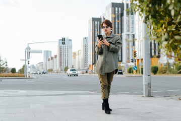 Fashionable young asian woman in stylish glasses holding a smartphone and looking at screen while standing on sidewalk in city. Slim woman using a mobile phone on street