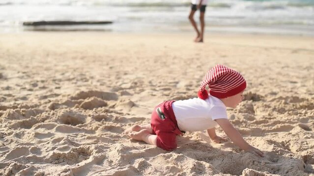 Little child playing on beach. Happy and joyful kid moves fast on all fours.  Kid in red-white striped hat, white t-shirt and red pants likes to crawl on warm sand.