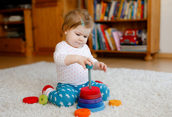 Adorable cute beautiful little baby girl playing with educational wooden toys at home or nursery. Toddler with colorful stack pyramid and music toy. Happy healthy child having fun with different toys
